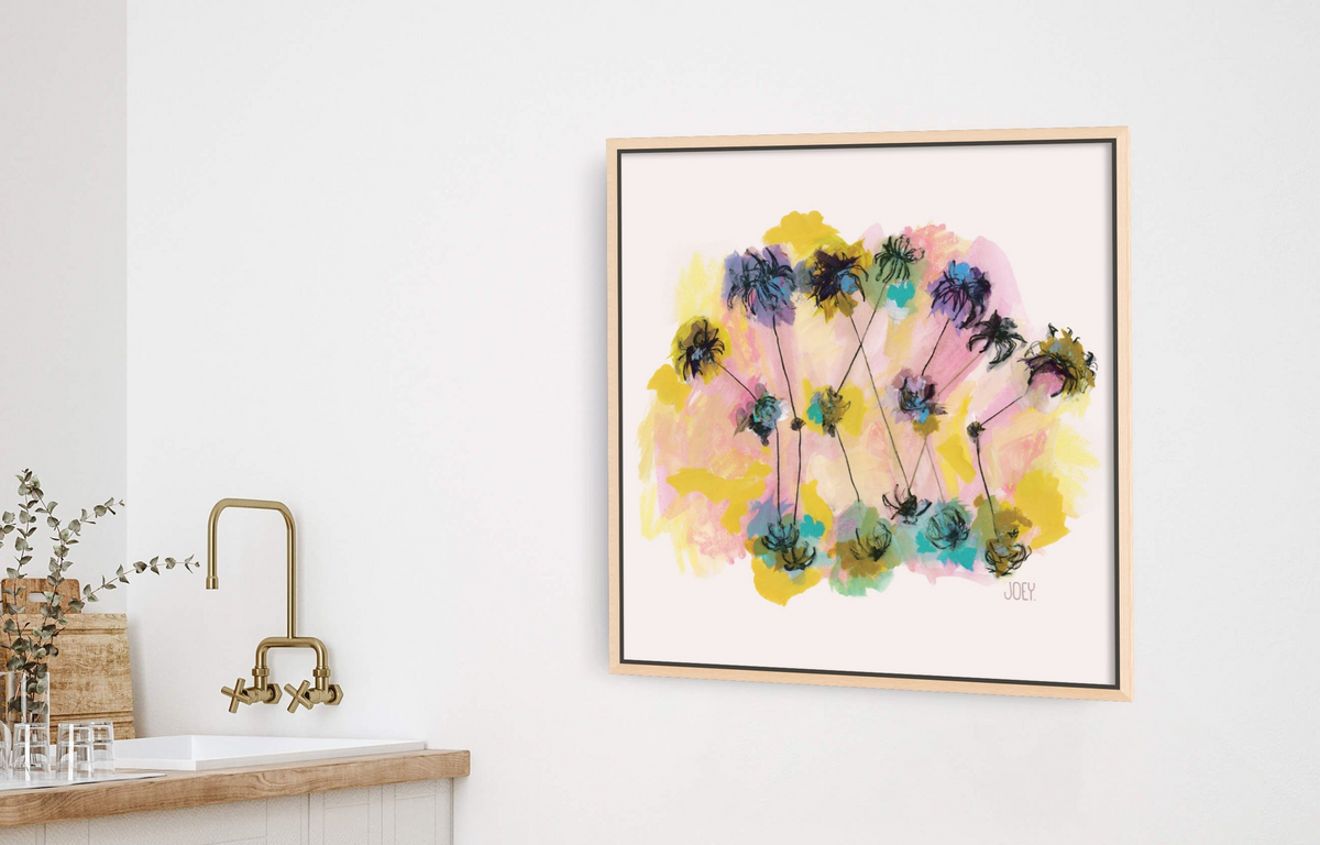 A framed abstract floral art print hangs on a white wall in a bright kitchen setting, with a sink, brass faucet, and two wooden stools visible below.